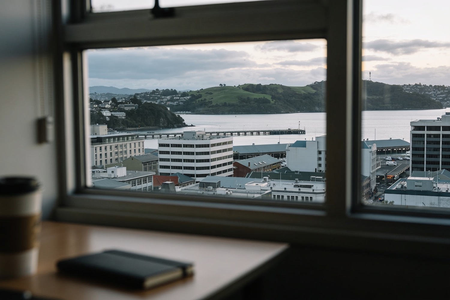 Looking out an office window at low-rise buildings, a harbour edge and green hills.